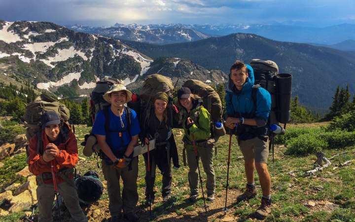 Five students with backpacks smile with a mountainous landscape in the background